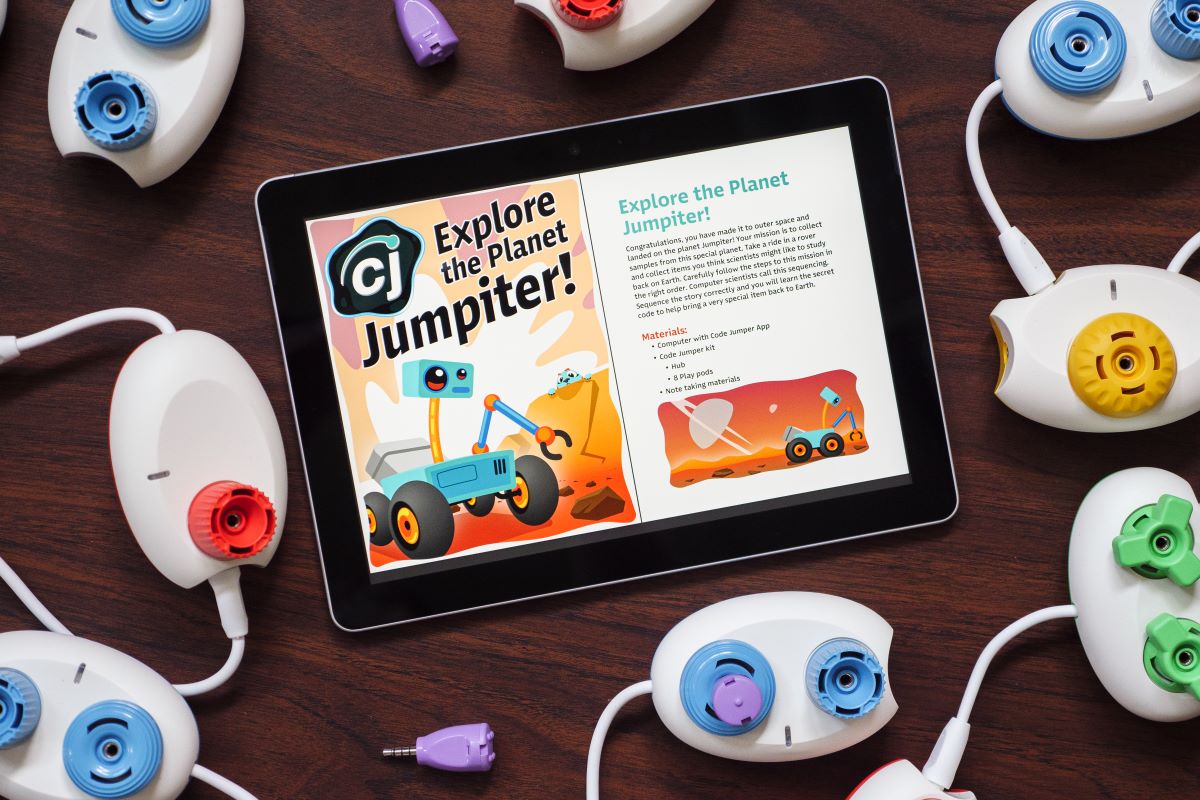 A tablet sitting on a table displaying the Code Jumper Puzzle — Explore the Planet Jumpiter! On the tablet's screen is an image of CJ, a blue cat-like creature on the surface of the planet Jumpiter observing a robot from behind. Code Jumper pods lay on the table surrounding the tablet.